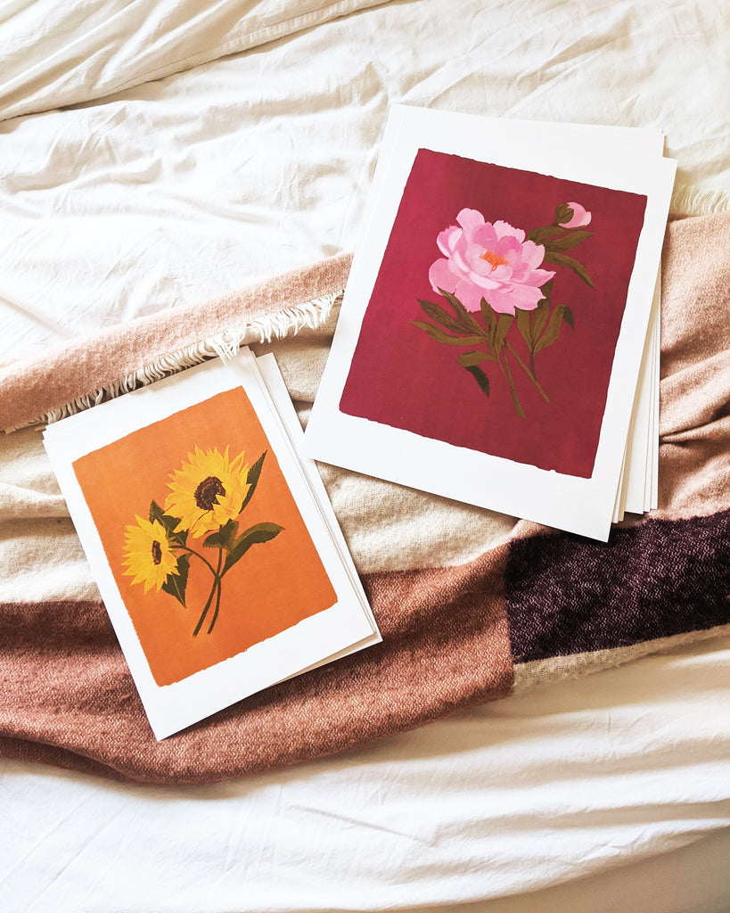 a stack of prints from the spring florals collection in A3 and a4 resting on a blanket as they wait to be hung. The designs on the very top feature a pair of Sunflowers on a burnt orange background, and a fluffy pink peony and flowerbud against a deep pink background.