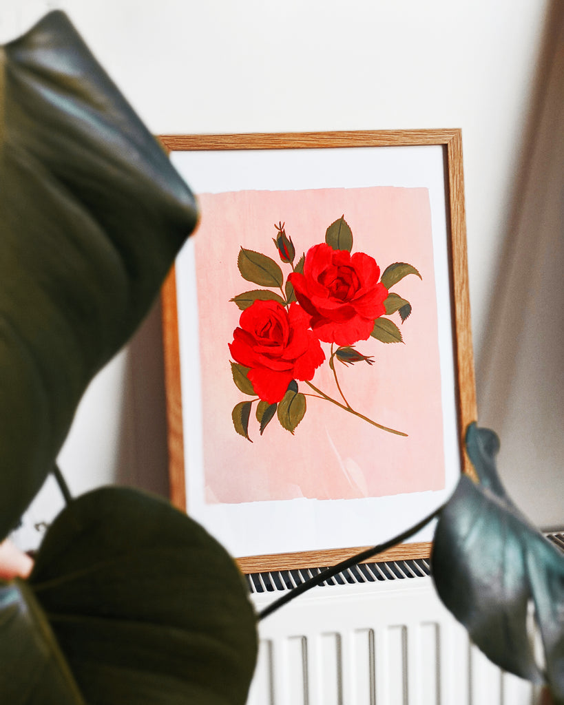 a pink and red print of painted roses rests, framed, ontop of a radiator, seen through the leaves of a houseplant