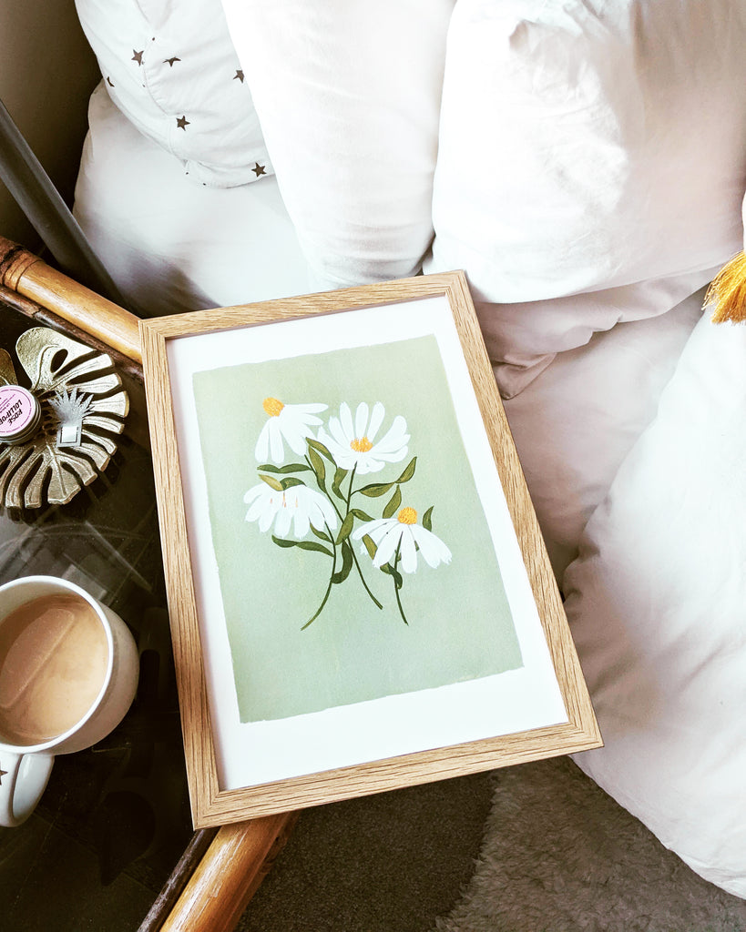 A framed print of the green and white coneflower print, resting on a bedside table next to a cup of tea 