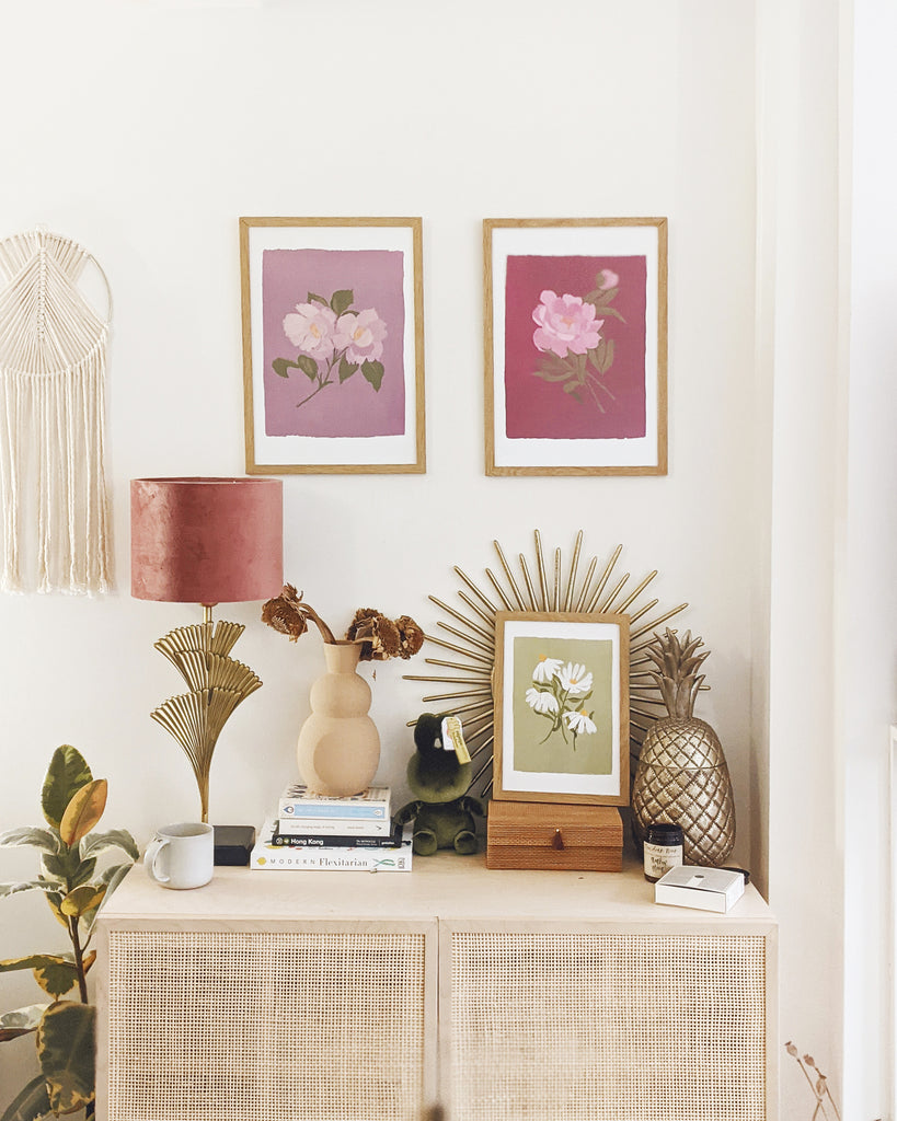 two floral prints - one deep pink and one pale lilac - framed, hanging above a rattan cabinet, decorated with al lamp, books, a vase of dried flowers and a framed green floral print