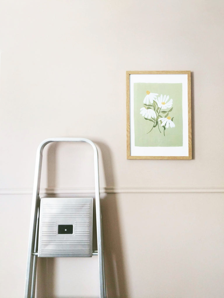 a green floral print in a pale wood frame, hanging on a pale lilac wall, next to a set of stepladders