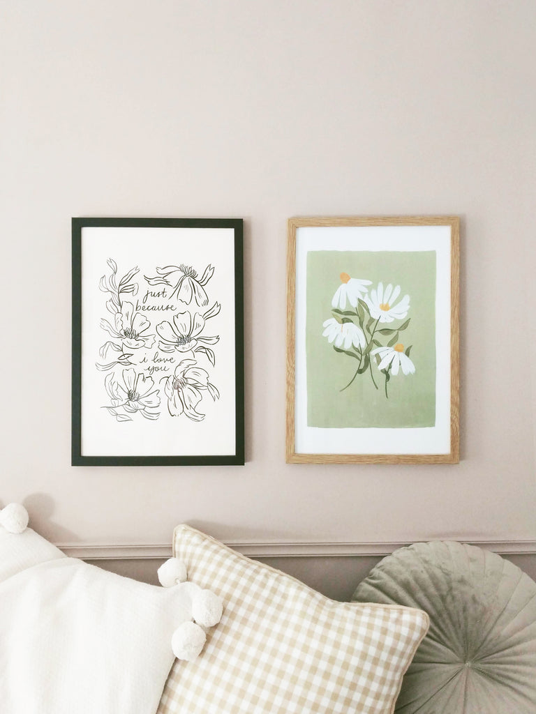 two prints - one black outline floral illustration, and one light green floral illustration, in frames, hanging on a pale lilac wall above a sofa filled with cushions