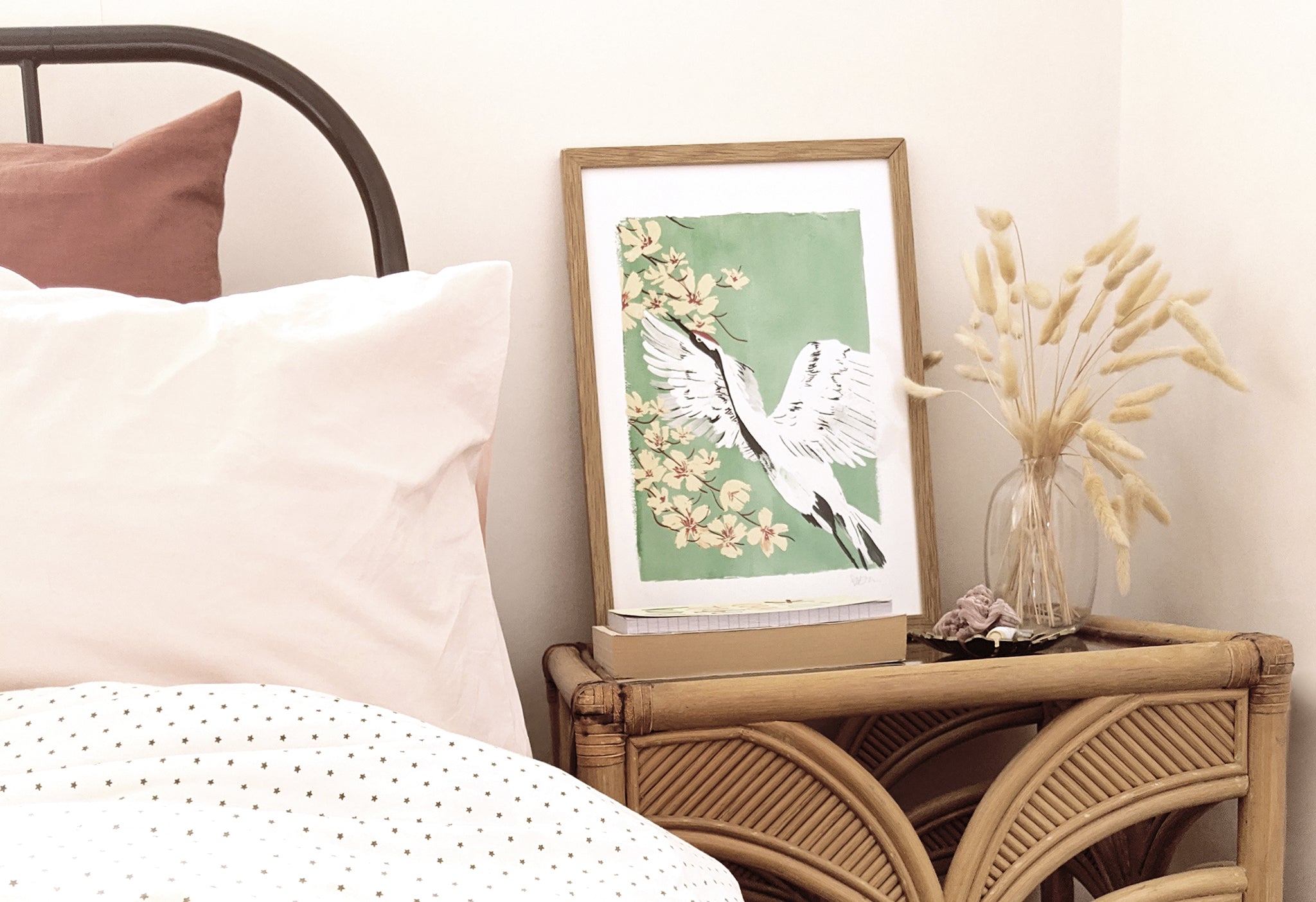 a beautiful print of a painted crane bird, soaring across a backdrop of pink blossom against a teal background, sits in an oak frame on a chic bedside table