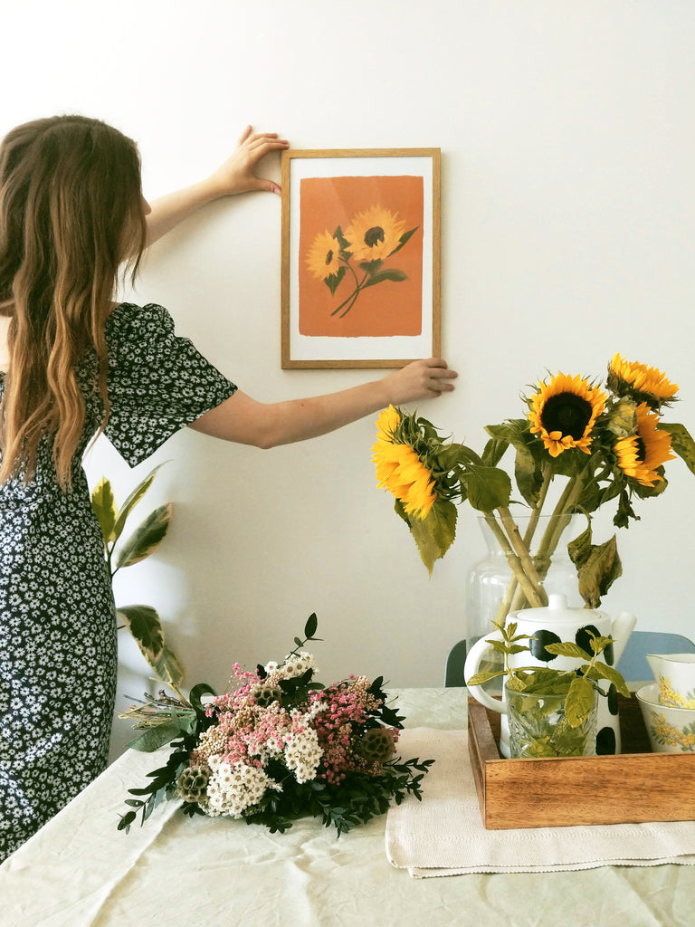 Annie, wearing a floral dress, hanging a sunflower print in a wooden frame, above a table with dried flowers and a vase of sunflowers.