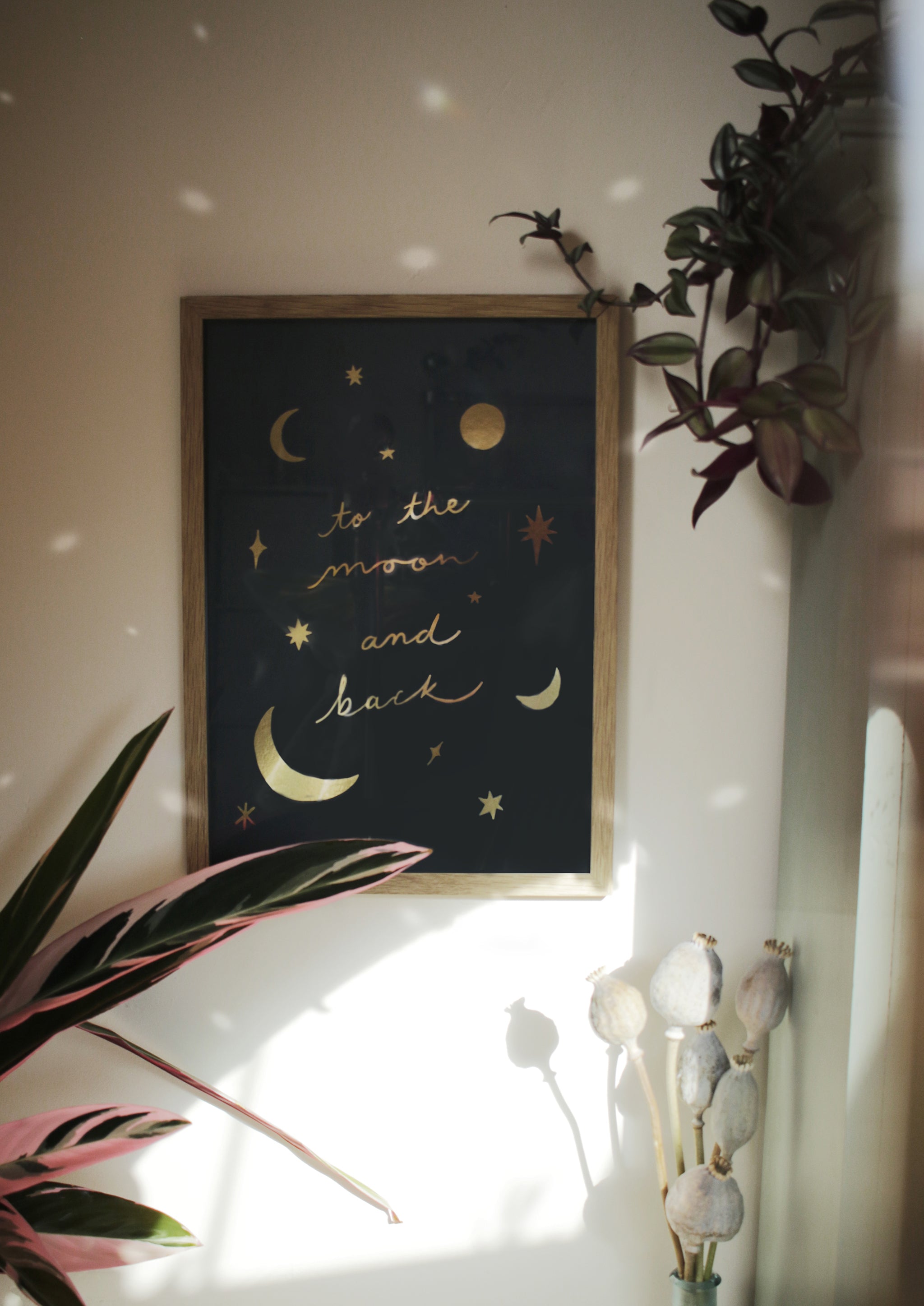 A glittering, gold foil print reads "to the moon and back" surrounded by gold stars and moons, hanging in an oak frame on a white wall, splashed in sunlight