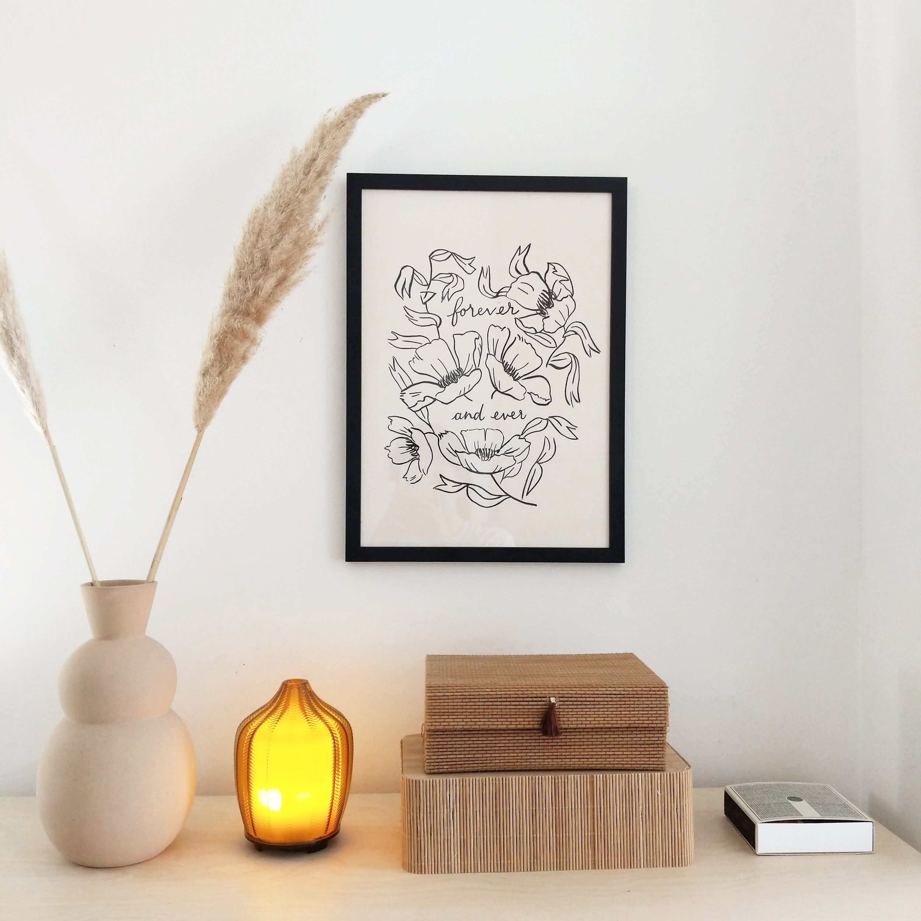A print featuring black, lineart-style flowers surrounding lettering reads "forever and ever", sitting in a black frame above a natural wood cabinet decorated with scandi-inspired boxes, vases and pampas grass. 