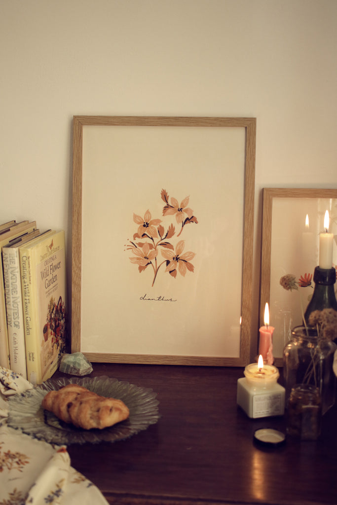 a print of a pink flower in a wooden frame, styled on a table alongside vintage books and candelight