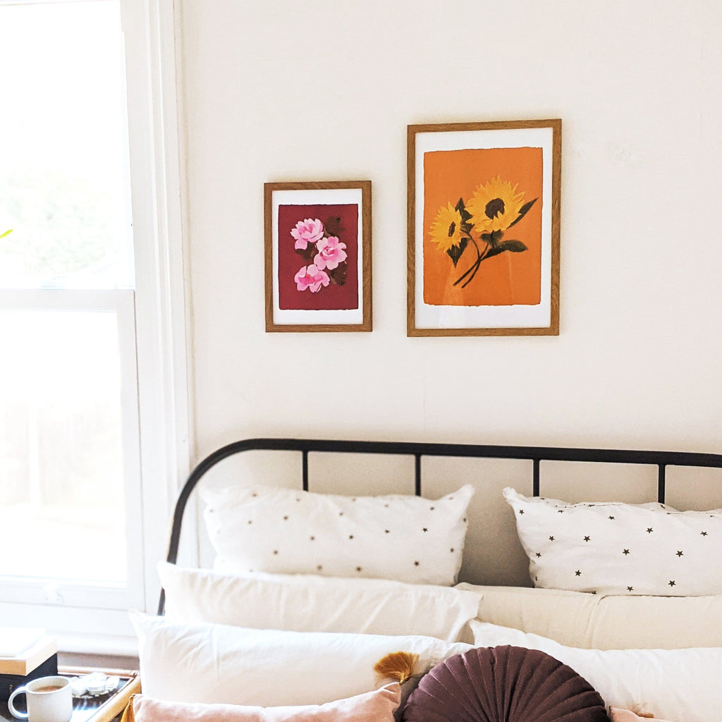 a light and airy bedroom with a cup of coffee on the nighstand. Two flroals printsh ang above the bed.