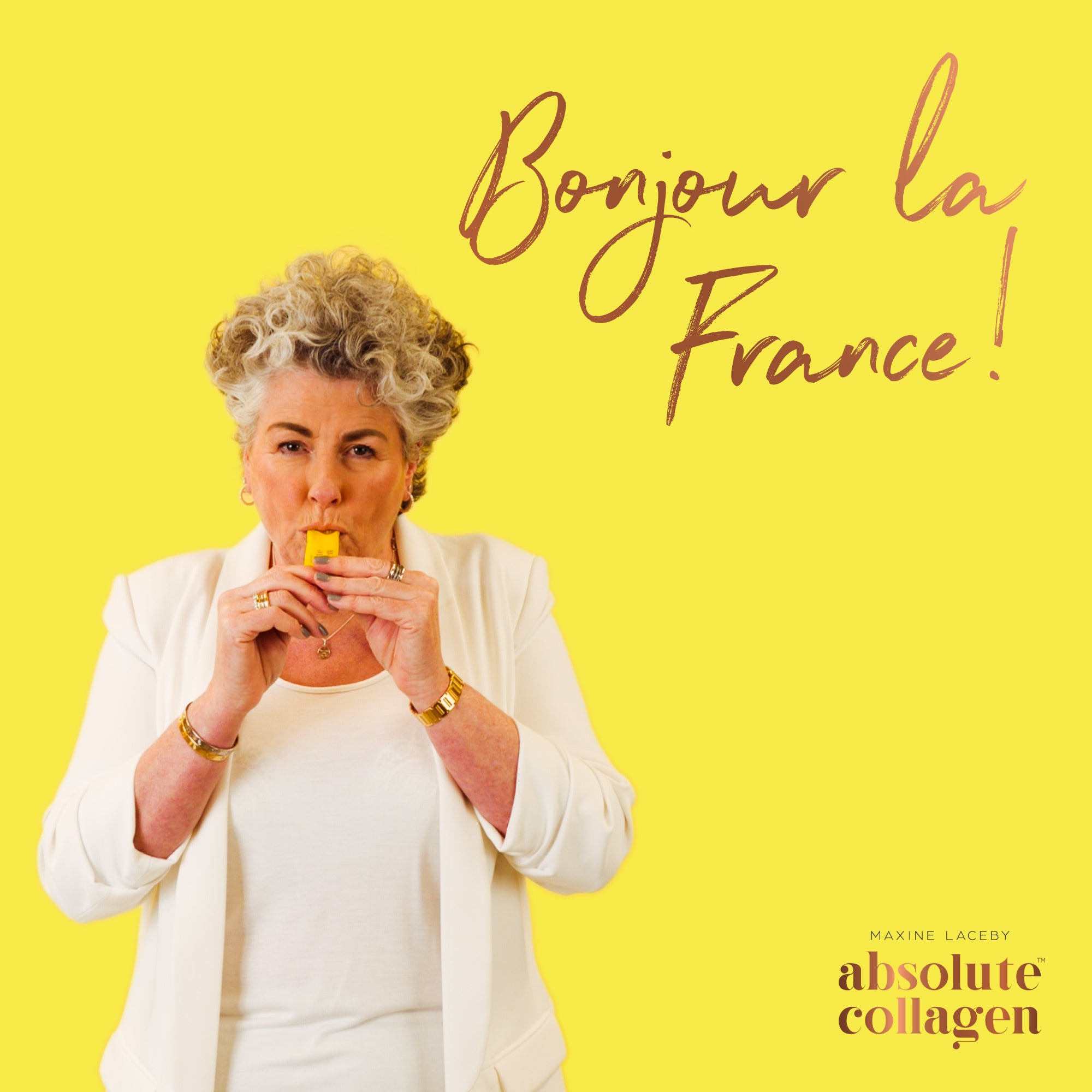 Photo showing Maxine Laceby wearing white and standing against a yellow background, she is taking a sachet of Absolute Collagen and the words Binjiur la France are emblazoned in copper writing to her right