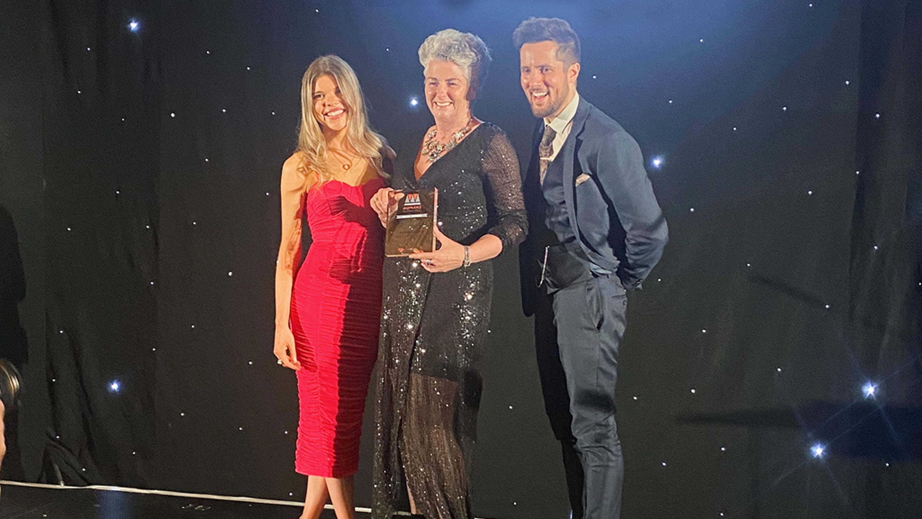 Photo showing Maxine Laceby, a smiling white woman with short grey hair, wearing a sparkly black outfit and posing with an award and two smiling presenters, a man and a woman