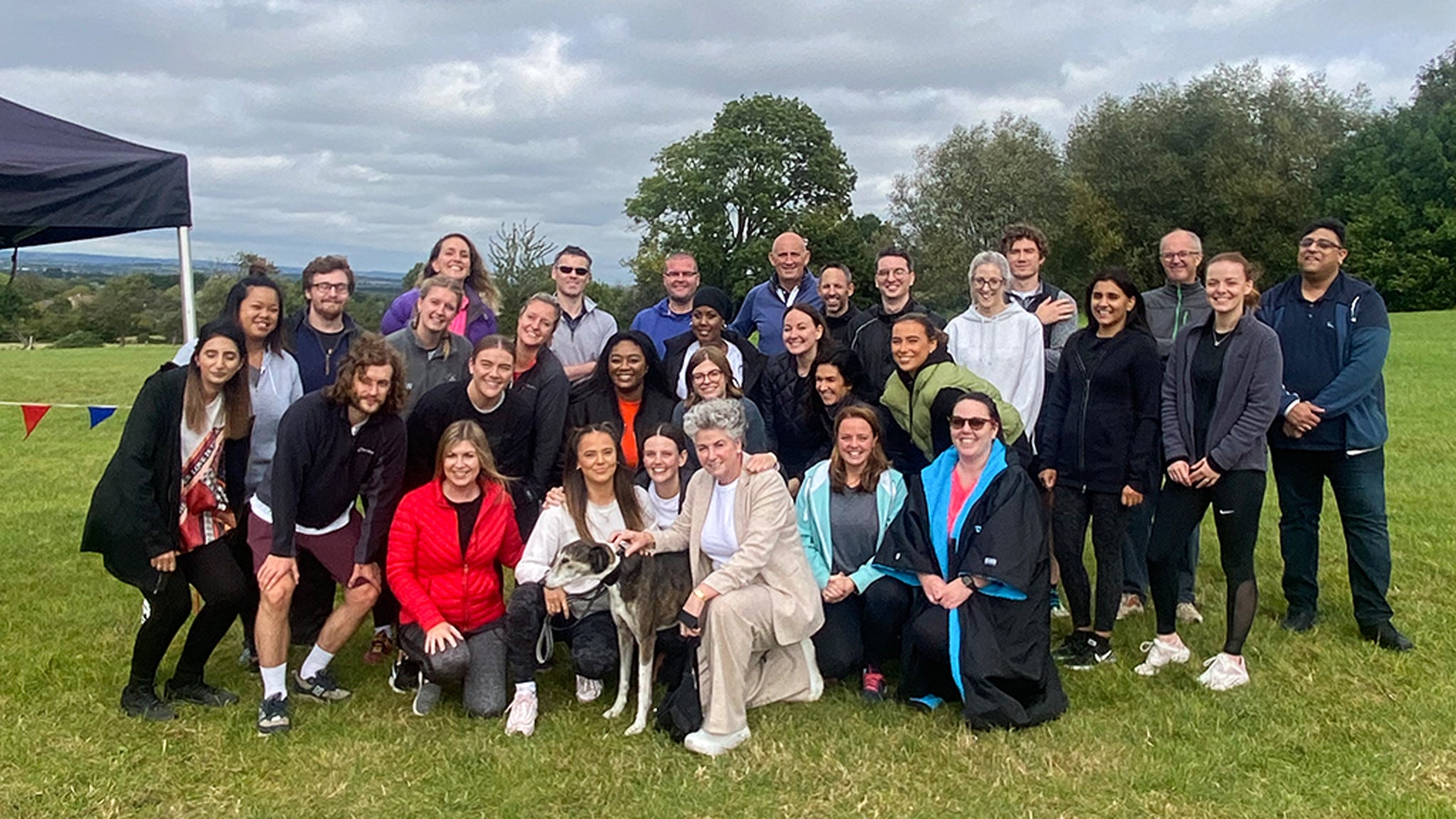 Photo showing a large group of people all wearing sporty outdoor clothes and standing together outside in the Costwolds, against a grey cloudy sky