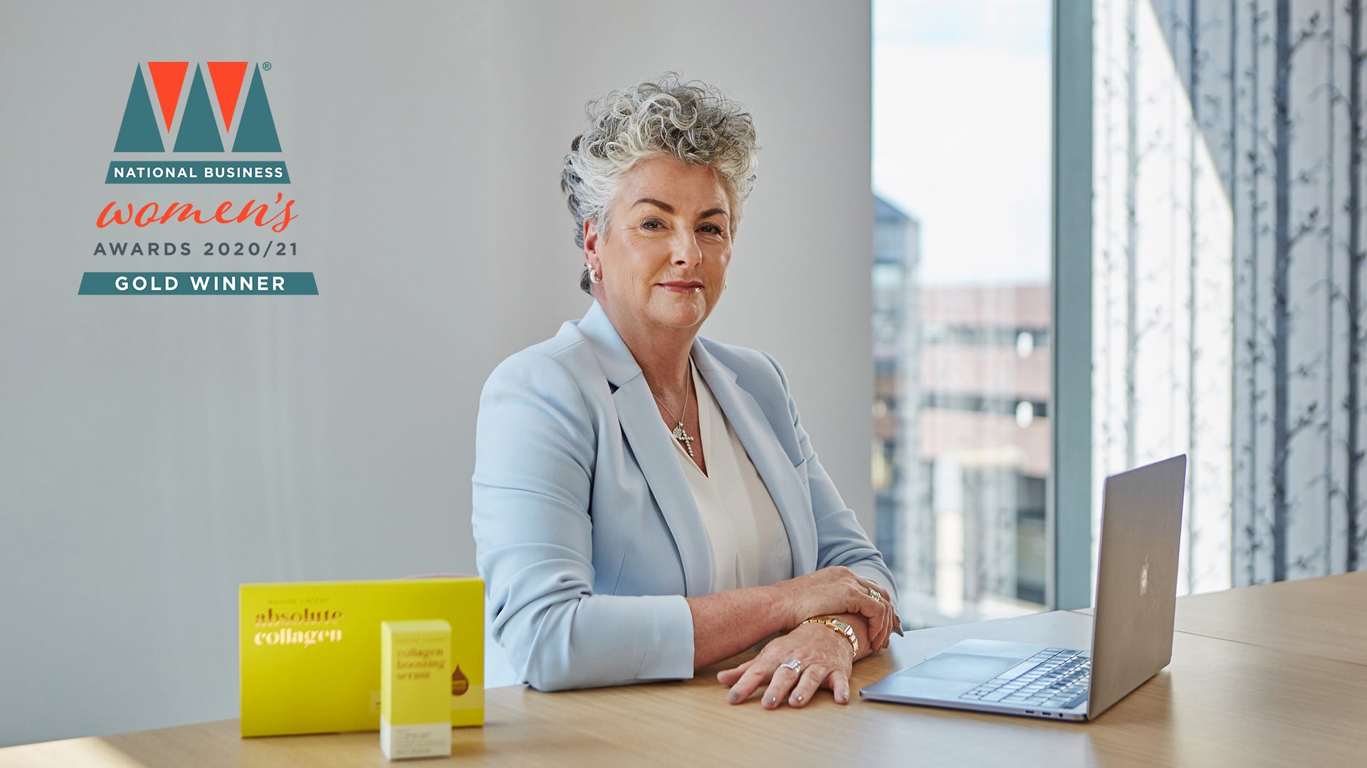 Photo showing Maxine Laceby wearing a pale blue blazer and sitting at a desk, she is looking at the camera and the National Business Woman Gold Winner logo is superimposed on the image to the left of her