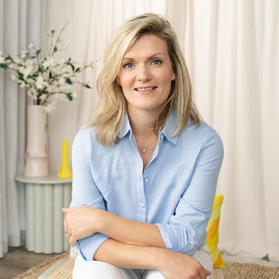 Woman in a blue shirt smiling, seated in a softly decorated room.