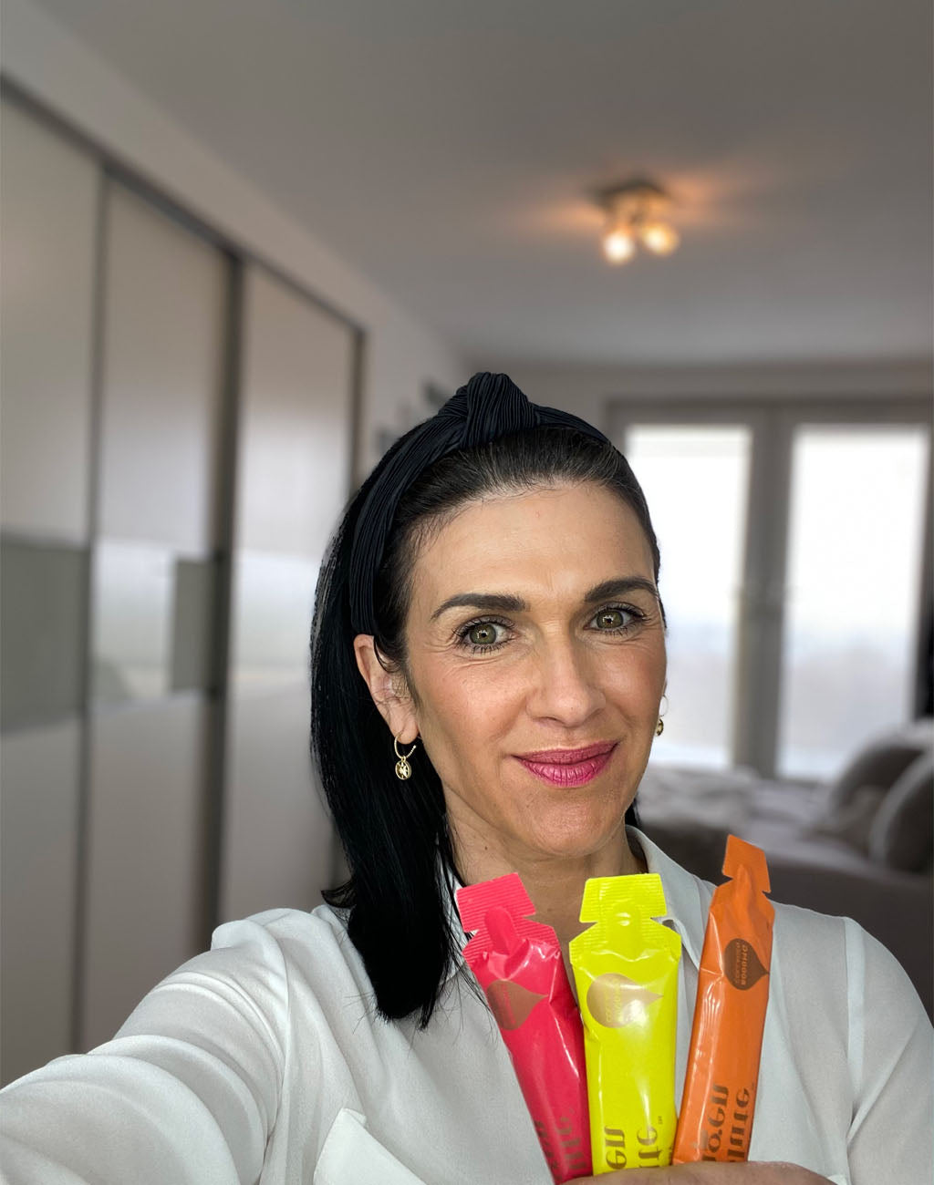 Photo showing a slightly smiling white woman with long dark hair, she is holding up three sachets of Absolute Collagen and taking a selfie in her bedroom