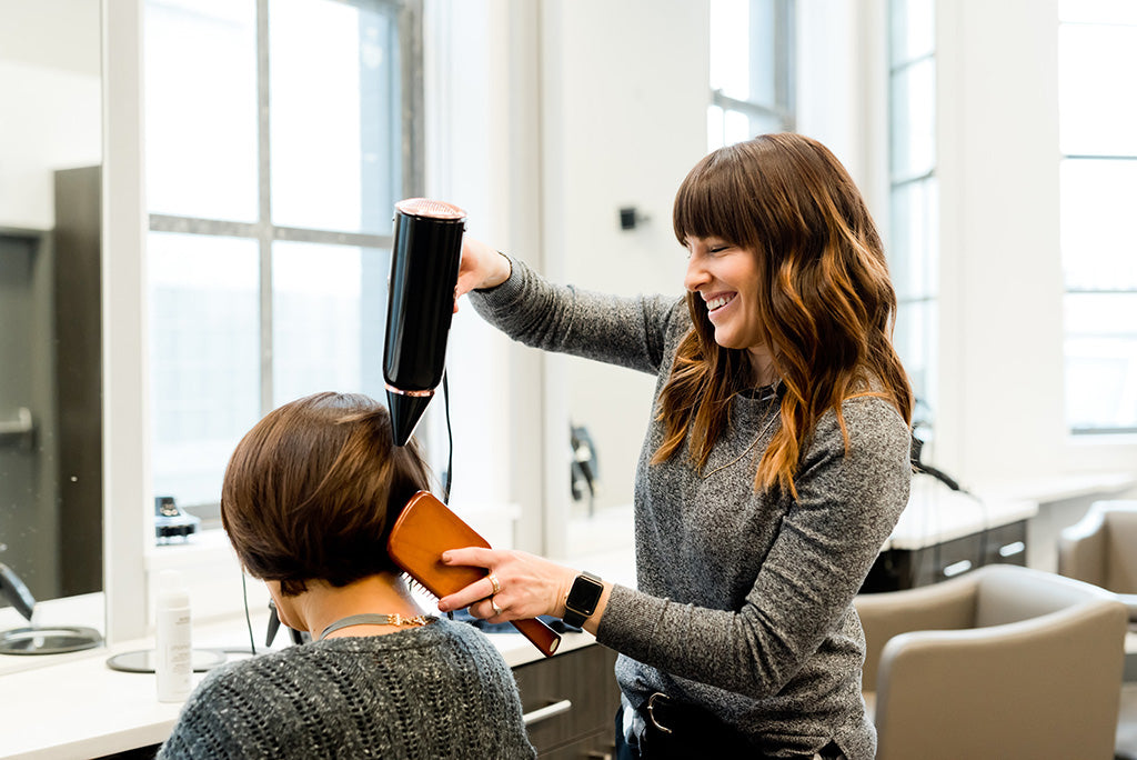 Photograph of a white woman holding a hairdryer and laughing with a hairdressing client sat in a chair with their back to the camera