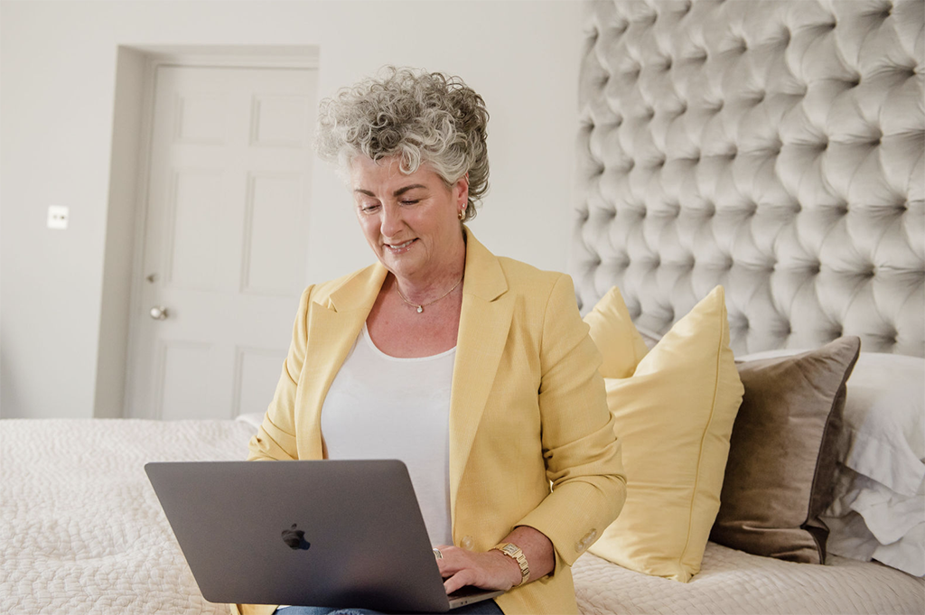 Maxine Laceby wearing a yellow blazer and sat on a bed working on a laptop