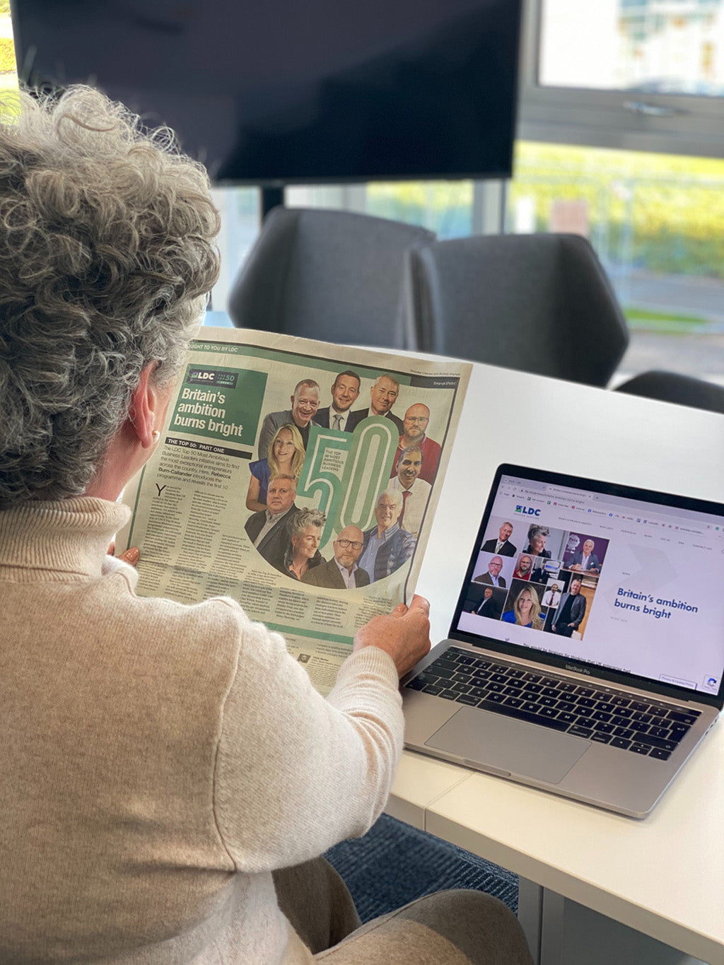 Photo showing a white woman with short silver hair sat at a laptop and holding a newspaper with her back to the camera