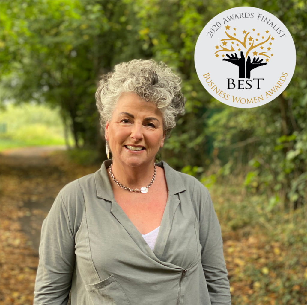 Photo showing a white woman with short wavy silver hair and wearing a grey top smiling at the camera against a forest background, with a roundel listing her as a Best Businesswoman Awards finalist in the upper left corner of the image