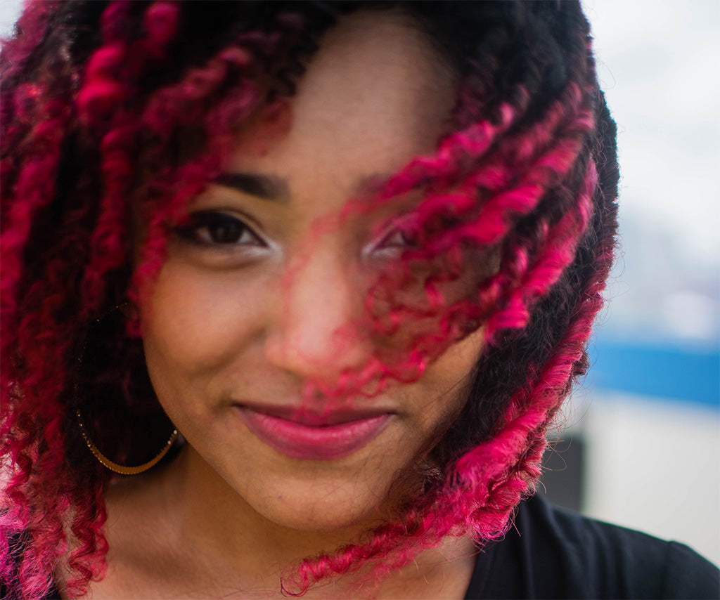 Photograph of a light skinned Black woman smiling into the camera with dyed pink hair blowing across her face