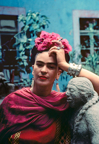 Frida Kahlo leaning against statue with magenta flowers in her hair wearing a matching rebozo
