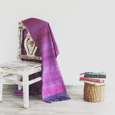 White chair with striped purple rebozo draped over with a basket of books beside it.