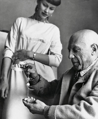 A black and white photo of Vivianna Torun Bülow-Hübe standing above seated Pablo Picasso who is holding a piece of her jewelry in his hands