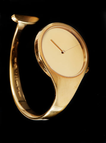 A golden wristwatch designed by Vivianna Torun Bülow-Hübe without an hour hand or numbers sitting against a black background