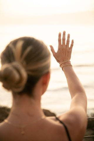Blonde woman wearing her hair in a low bun holds her right hand up to the sun wearing gold bracelets.