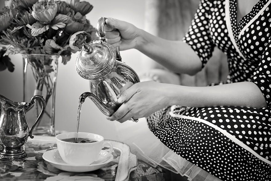 A greyscale image. A lady in a dark polka dot dress, sitting, pouring tea from a silver teapot