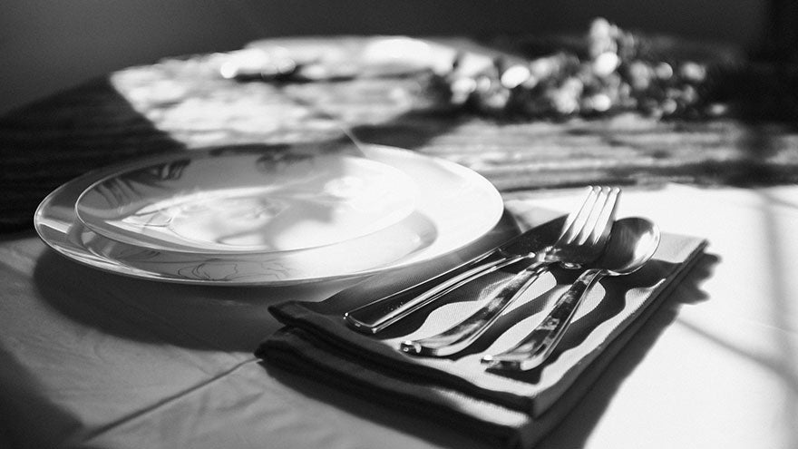 A table with a tablecloth, upon which there is a knife, fork, and spoon lying on a folded napkin to the right of a gleaming plate