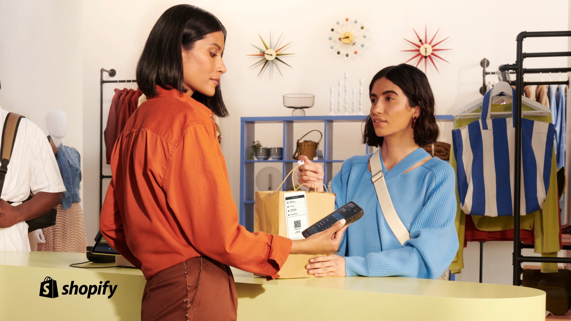 Woman in blue sweater at a check out counter taking her bag as another woman in an orange shirt looks at her handheld POS 