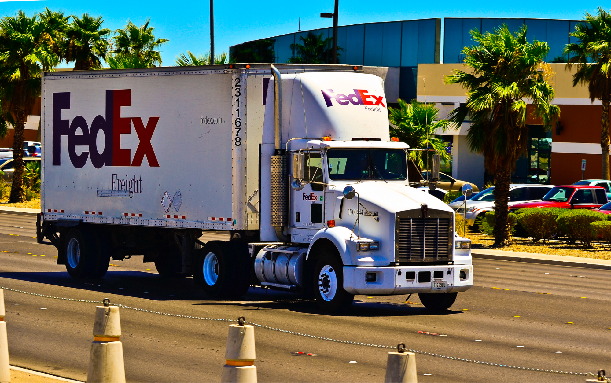 A FedEx freight truck driving down a highway bordered by palm trees on a Sunday day.