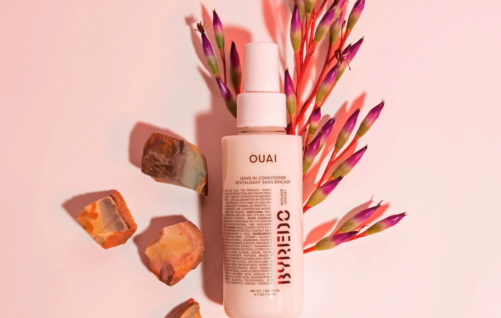 Pink OUAI and BYREDO leave-in conditioner bottle on purple and green flowers and a pink background