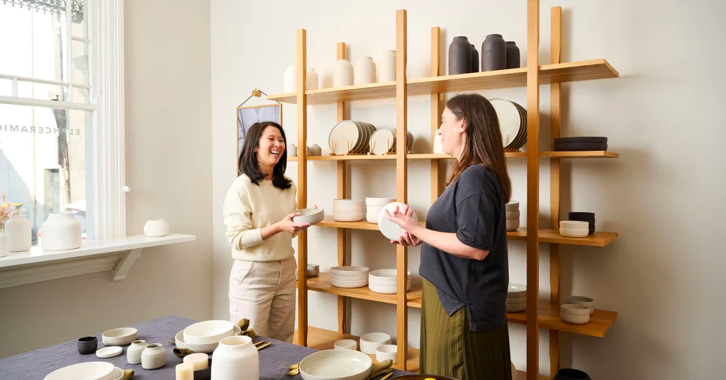 In Elph Ceramics' storefront, simple wood shelving holds pottery, with owners standing in front.