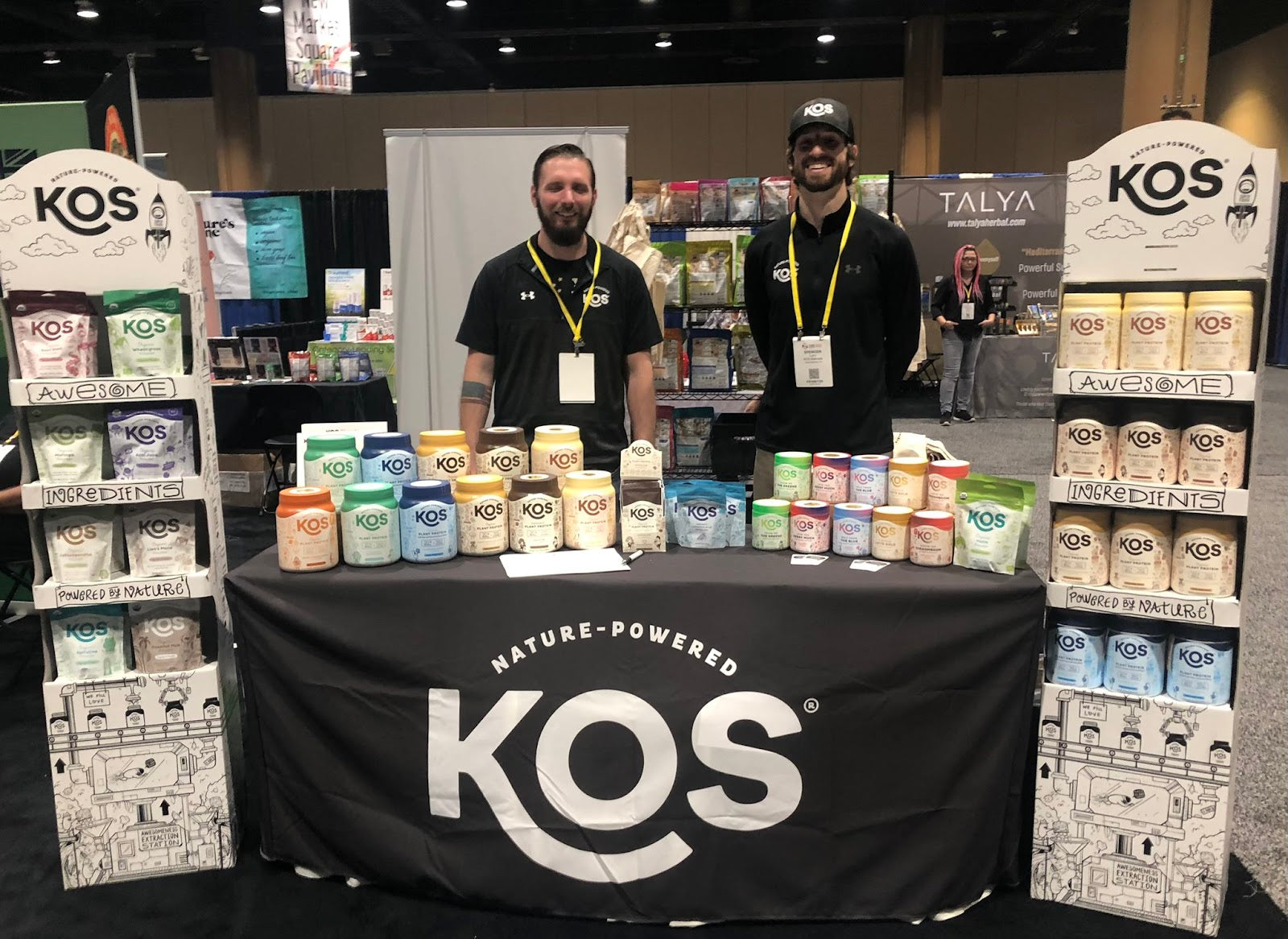 Two men stood behind a black table with KOS in large letters. The booth is enclosed by two high-standing displays with packaged protein powder.