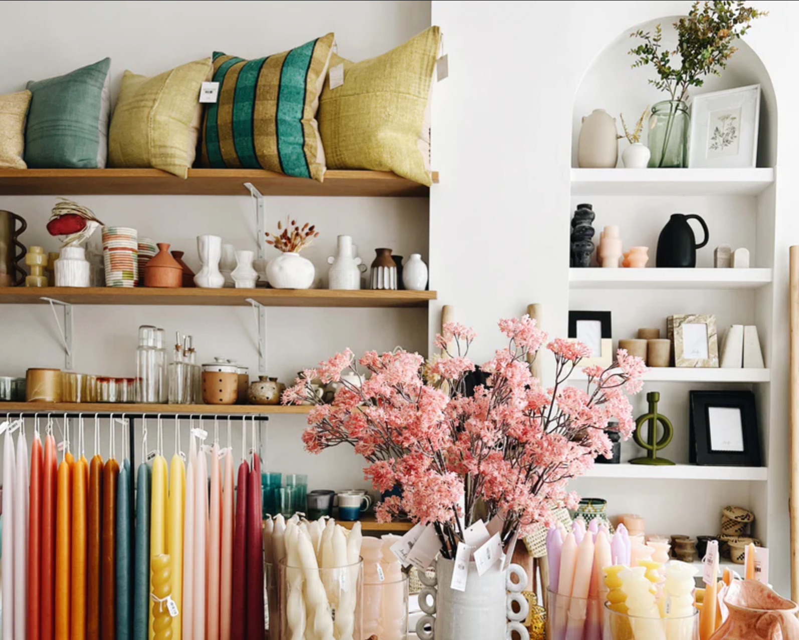 Cushions, vases, and other home decor objects line wooden shelves with candles and flowers in the foreground.