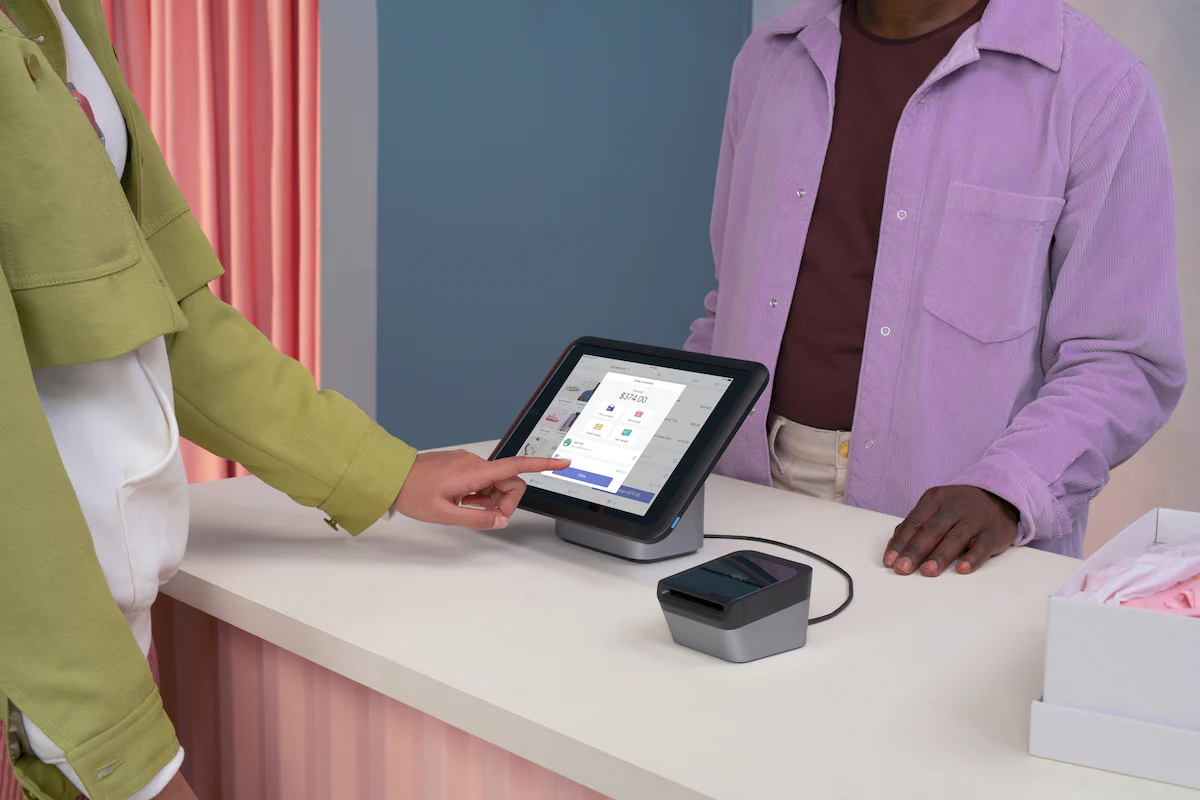 Photo of two people at a checkout desk. The shopper is tapping the POS tablet.