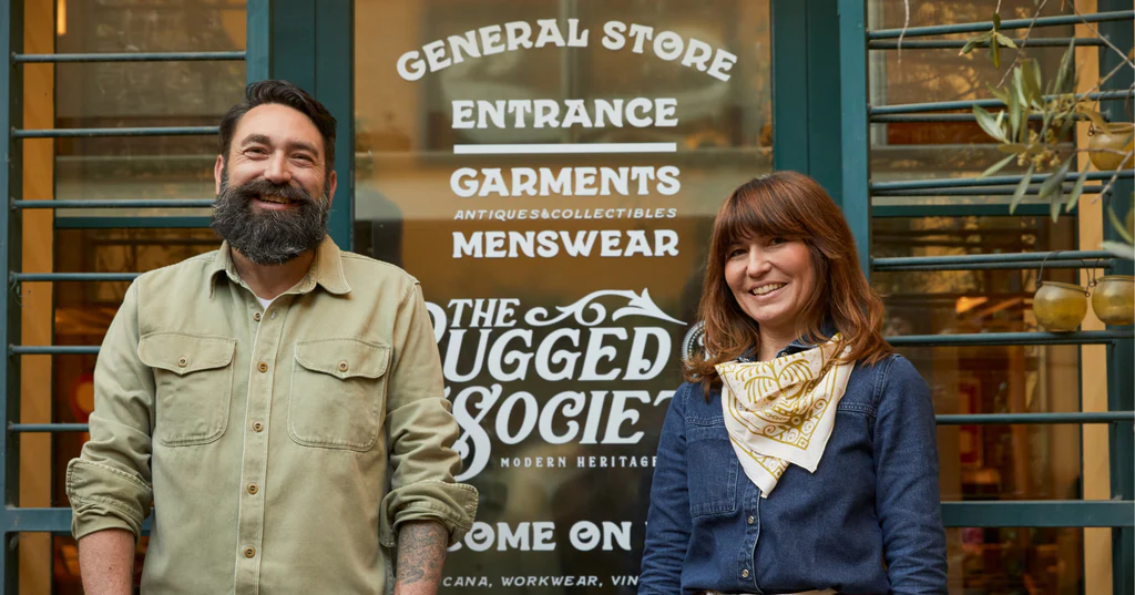 The Rugged Society's front window with hand-lettered decals and owners standing in front.