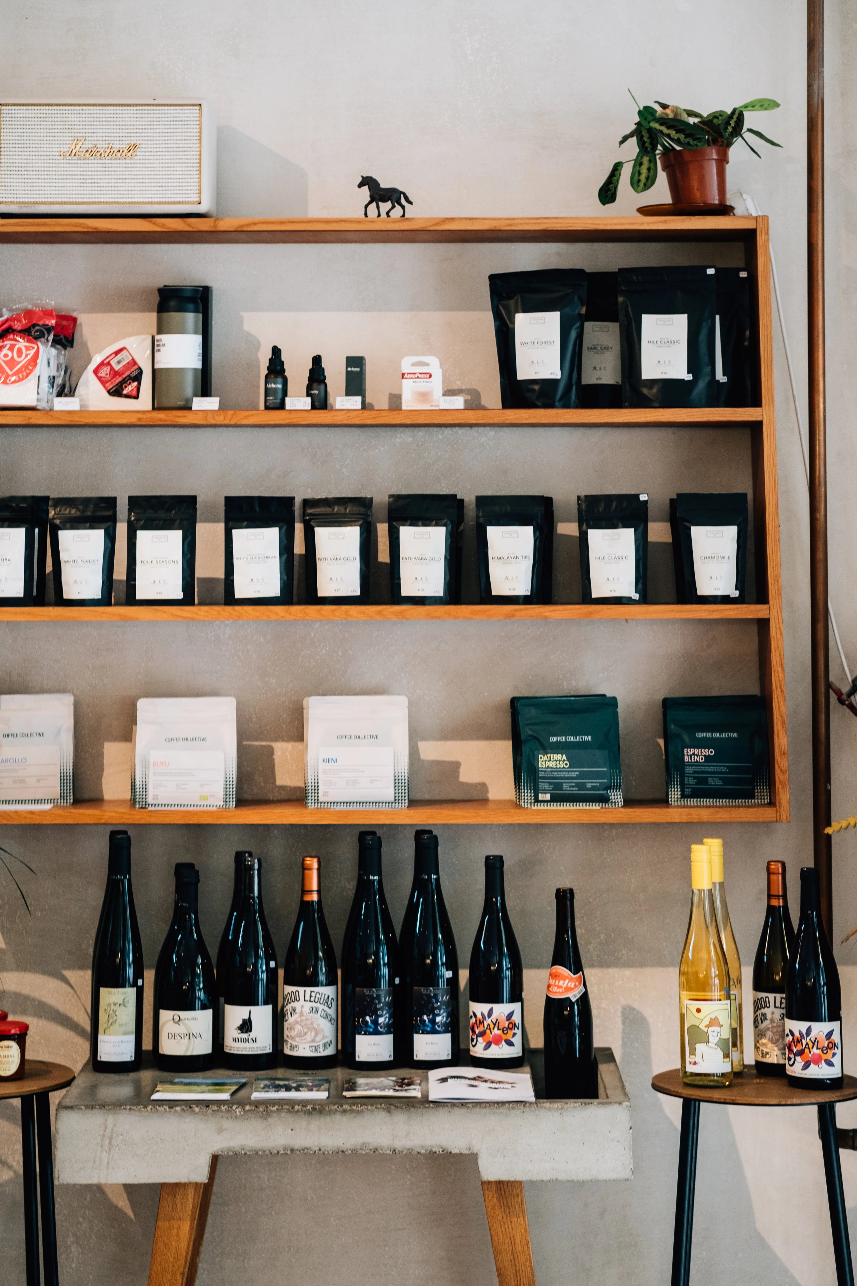 Wooden shelf holds black or white small pouch bags. Glass wine bottles with a variety of decorative labels line the bottom on a grey cement shelf and small wooden tables.