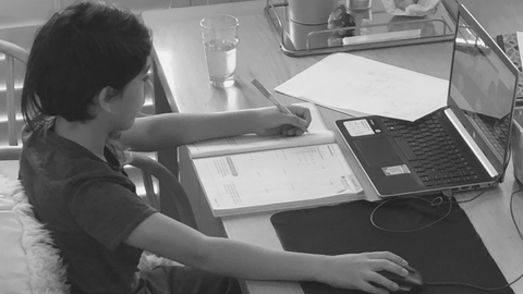 Child boy sitting at a table looking at a laptop with study materials stacked to the right and left of keyboard.