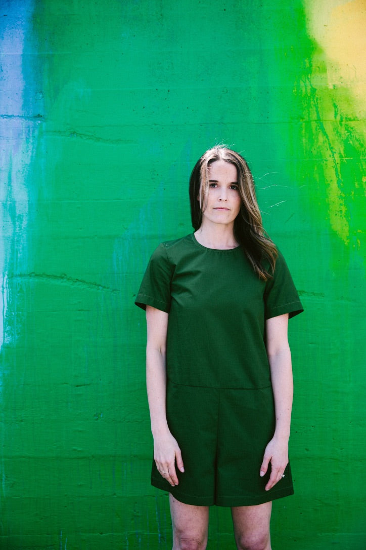 a white woman with brown hair wears a green romper in front of a green wall
