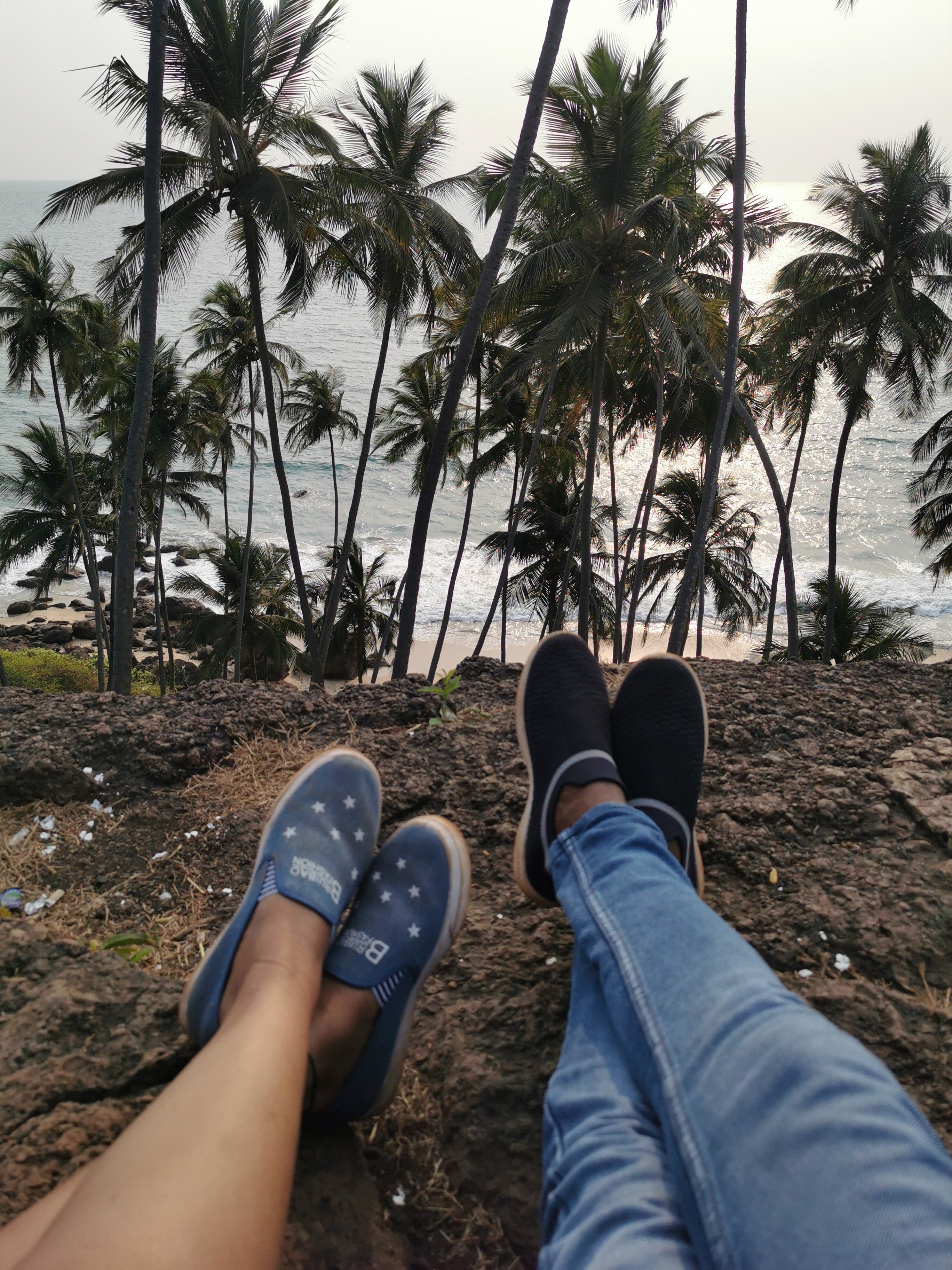 Two people sitting on a cliff by the beach wearing tennis shoes