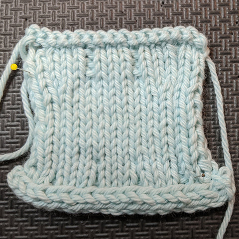 Photo of a knit square in a light blue yarn. The center of the swatch has stockinette with 3x1 ribbing on both ends. One end has an EZ sewn bind-off, the other has an i-cord bind-off. 