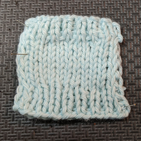 Photo of a knit square in a light blue yarn. The swatch has stockinette in the center and ribbed edging on the top and bottom. 