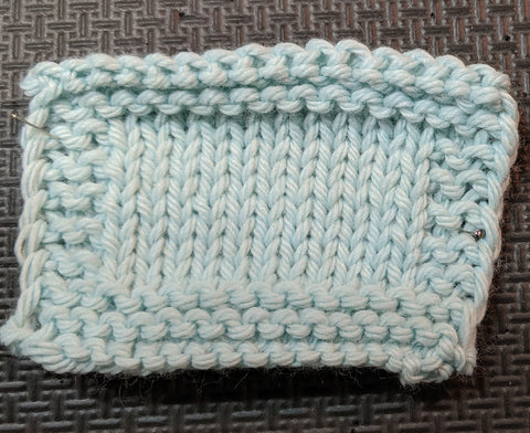 Photo of a knit square in a light blue yarn. Stockinette is in the center of the swatch, with garter stitch around all four edges. 