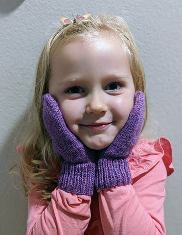 Photo of a young blond girl in a pink shirt wearing purple mittens on both hands, with her hands framing her face.