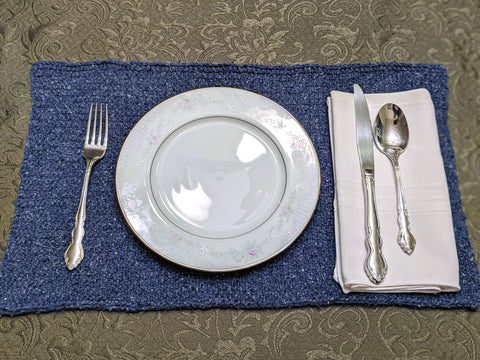 Photo of a table place setting. Brown floral table cloth with a denim blue colored knit placemat with a china plate and utensils.