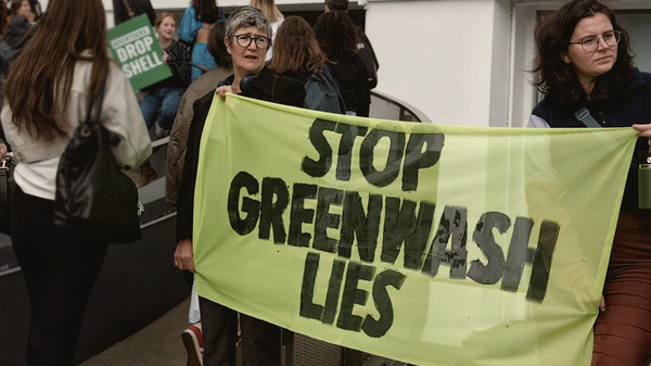 Two protestors hold up a green sign that protests greenwashing. In the background another protester holds up a poster demanding a major corporation stop protesting.