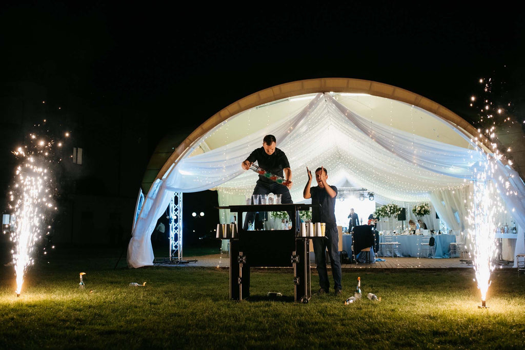 Two bartenders perform a show at a nighttime outdoor wedding.