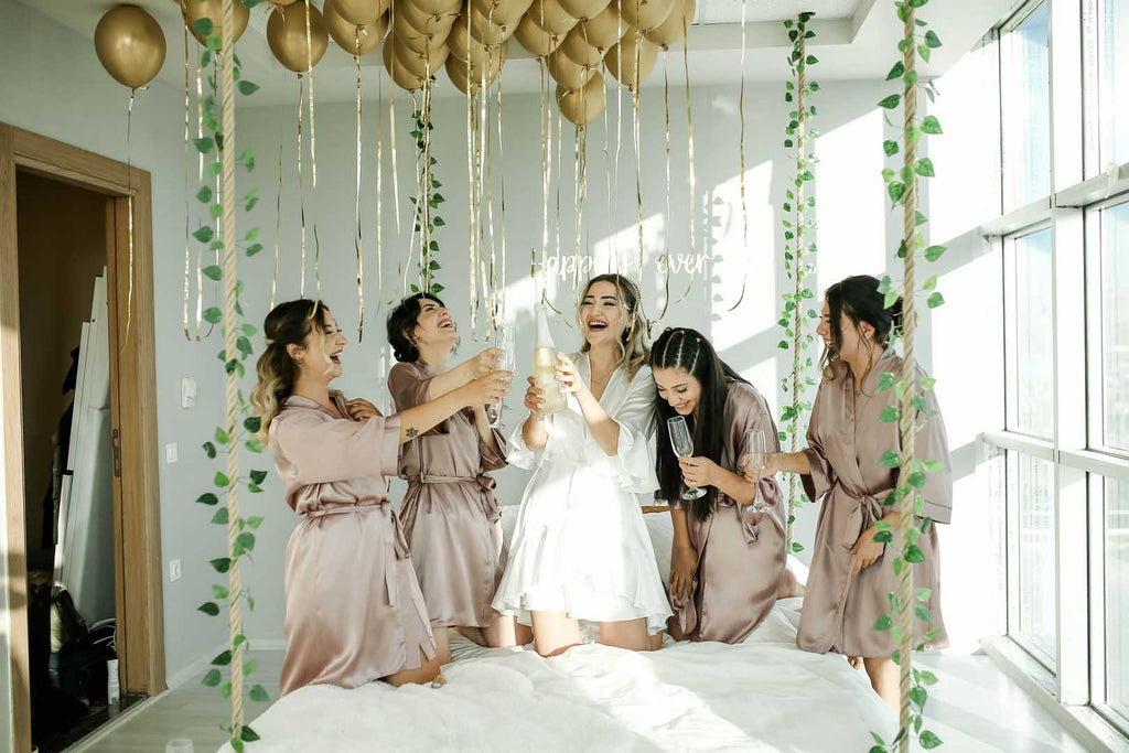 Four women in lavender silk bathrobes and one women in a white bathrobe prepare to cheers as the women in the white bathrobe opens a bottle of champagne. The room is decorated with green hanging vines and gold balloons.